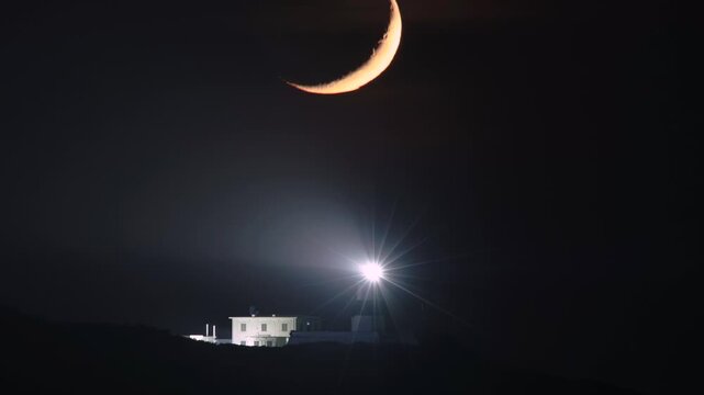 A glowing crescent moon hangs in the dark night sky above the bright flashing light of Fuguijiao Lighthouse in Shimen, New Taipei City, Taiwan.