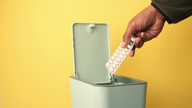 Person throwing away blister pack of medicine into small trash bin on yellow background