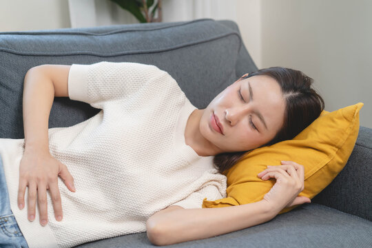 Close-up of a young woman sitting on the sofa and holding her stomach, suffering from stomach ache, abdominal pain or menstrual cramps at home.