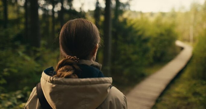 Rear side profile of a woman looking toward a wooden boardwalk through an autumn forest. Peaceful outdoor scene with natural daylight, woodland surroundings, and a calm hiking atmosphere.
