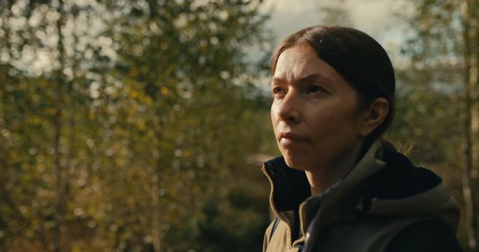 Portrait of a woman standing in an autumn woodland looking ahead with a focused, thoughtful expression. Natural outdoor setting with soft daylight, shallow depth of field, and seasonal foliage.