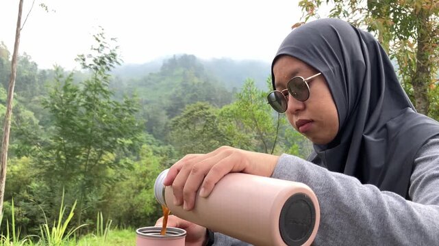 Slow motion clip of a young Muslim woman wearing a hijab and sunglasses pouring hot coffee from a thermos into a cup while relaxing in a beautiful misty green mountain forest. Outdoor camping concept.