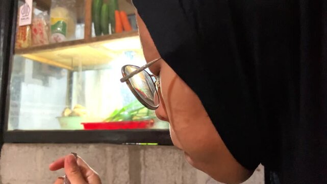 Slow motion close-up shot of a young Muslim woman wearing a black hijab and glasses enjoying a meal of fried noodles using a spoon at a traditional local food stall or restaurant.