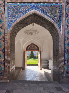 Ornate tiled archway at Shah-i-Zinda necropolis in Samarkand with carved wooden doors opening to a green courtyard. Ideal for travel, heritage, and cultural publications.