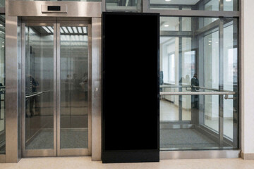 Naklejka na ściany i meble Black blank vertical led screen mockup standing beside metallic elevator doors in modern office lobby. Empty digital signage board for advertising displays information inside glass building interior.