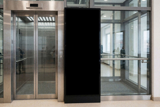 Black blank vertical led screen mockup standing beside metallic elevator doors in modern office lobby. Empty digital signage board for advertising displays information inside glass building interior.
