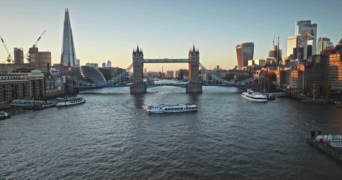 Aerial panorama of London cityscape at sunset, iconic Tower Bridge spanning the River Thames with tourist boats sailing below, and The Shard dominating the modern skyline under a warm evening glow