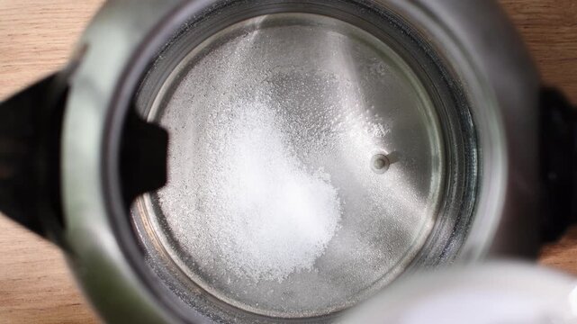 Person pours three spoonfuls of citric acid into electric kettle to remove mineral scale during routine kitchen cleaning.