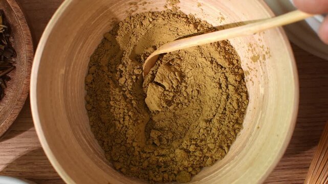 Hand stirs roasted hojicha powder with bamboo spoon in wooden bowl, showing fine texture during tea preparation ritual.
