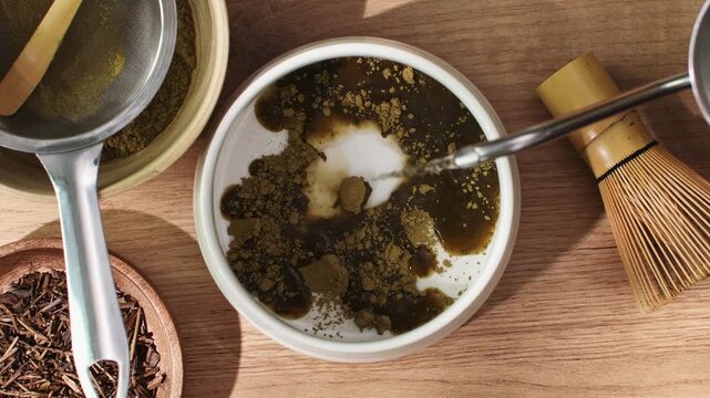 Hand pours hot water from gooseneck kettle over sifted hojicha powder in bowl to begin traditional Japanese tea preparation.