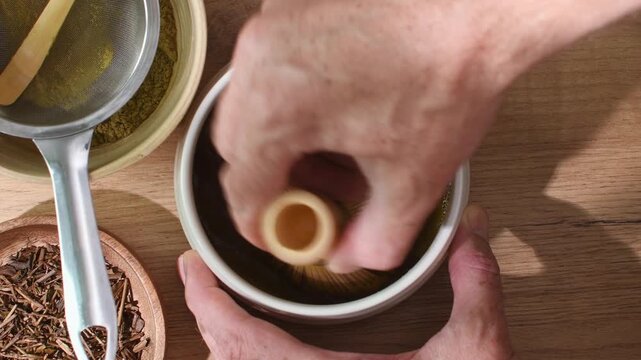 Hands whisk roasted hojicha powder with bamboo chasen in ceramic bowl to create smooth tea during traditional tea ritual.