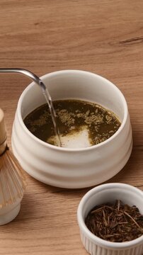Hot water pours from gooseneck kettle onto sifted hojicha powder in ceramic bowl during Japanese tea preparation ritual.
