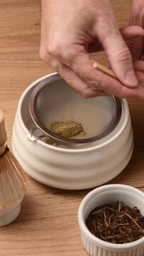 Hands sift roasted hojicha powder through fine mesh sieve with bamboo spoon into ceramic bowl for smooth tea preparation.