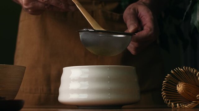 Hands sift roasted hojicha powder through fine mesh sieve into ceramic bowl during Japanese tea preparation step at home.