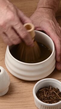 Hands whisk roasted hojicha powder with bamboo chasen in ceramic bowl during traditional Japanese tea preparation ritual.