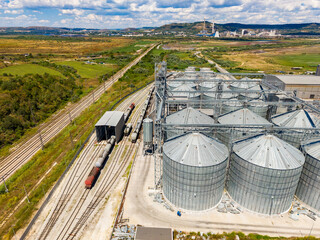 Storage silos and train tracks in industrial area with green fields and mountains in background under cloudy sky © DSMT