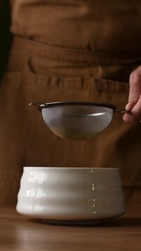 Hands add roasted hojicha powder to fine mesh sieve and sift it into ceramic bowl during Japanese tea preparation ritual.
