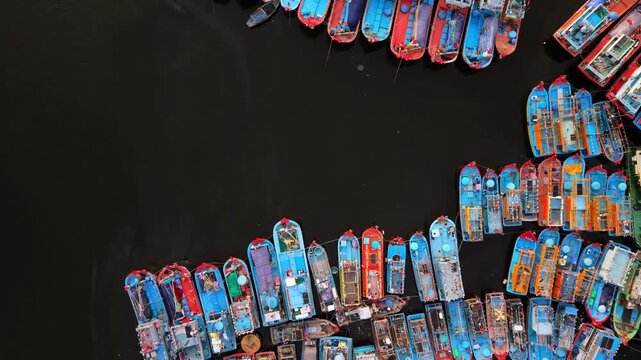 Vertical drone footage of colorful fishing boats moored side by side in a calm harbor, creating striking geometric patterns and vibrant maritime scenery.