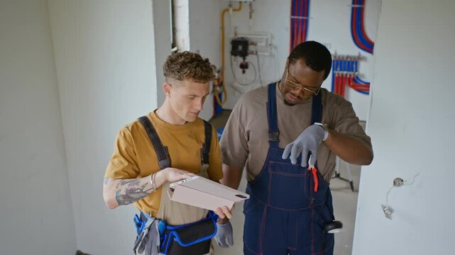 Caucasian and African American male builders discussing renovation plans on tablet computer in unfinished room. Perfect for construction, home improvement, and real estate marketing campaigns