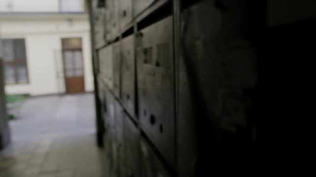 Tracking shot moving past rows of old, weathered mailboxes in a dark, neglected hallway. The camera reveals a glimpse of an old building courtyard through the open doors.
