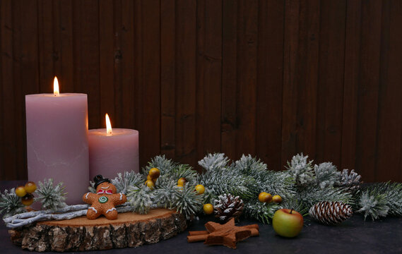 Christmas arrangement on a wood slice against a dark wooden background, featuring two lit candles, a gingerbread man, cinnamon sticks, an apple, and snow-dusted fir greenery, with space for text.
