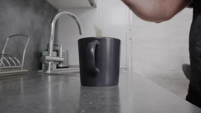 Man preparing tea in a kitchen, adding a teabag to a mug. Steps show pouring boiling water, steeping, and removing the bag. Routine beverage preparation in a home setting.
