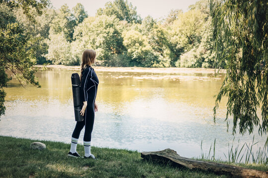 Woman with yoga mat standing by lake, contemplative nature walk, solo fitness journey, scenic workout motivation, peace and solitude