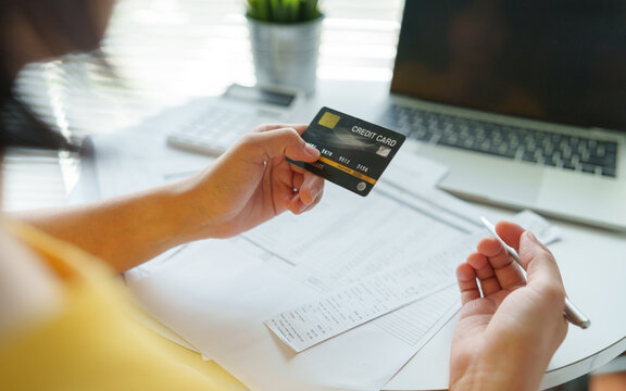 Asian Woman holding a credit card while reviewing bills and budget beside a laptop, representing online payment and personal finance management.