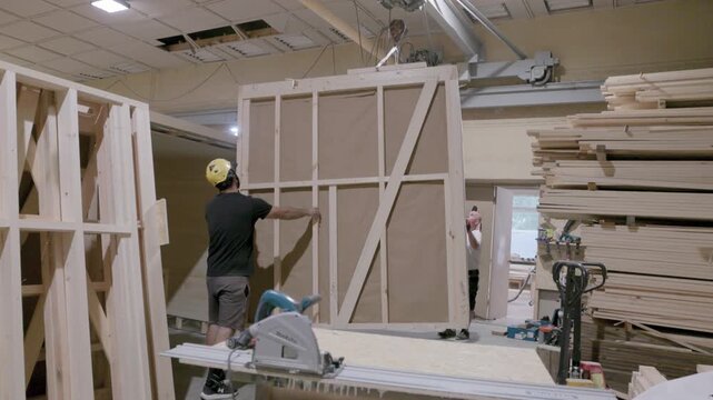 Factory worker wearing a safety helmet guides a suspended timber wall panel beside stacked lumber during prefabricated cabin production