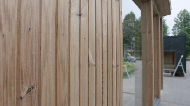 Close view of vertical timber cladding and large glass windows on a small wooden cabin, with reflections, trees, and construction activity in the background