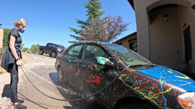Person washing car, in residential driveway, with car, water hose, soapy water, with nearby objects.