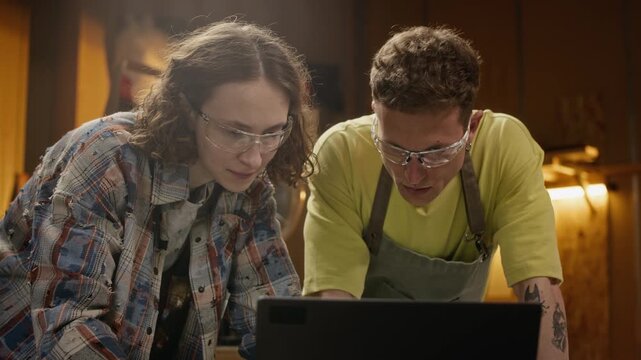Young adult Caucasian male and female carpenters wearing safety glasses looking at laptop in workshop. Video is ideal for small business marketing and vocational training ads