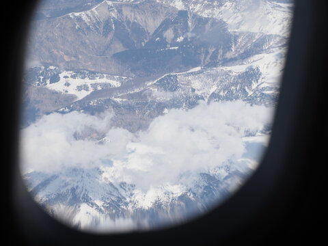 Panoramic view of snow covered mountains and clouds through a round aircraft window