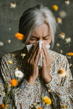 Woman with a wrinkled face shows discomfort while blowing her nose with a napkin. Flowers and pollen scatter in the background, highlighting her allergy