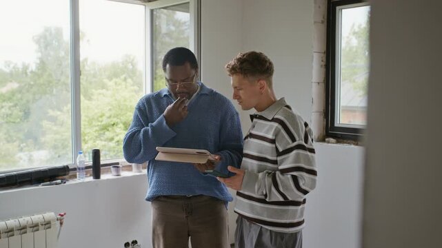 African American young adult and Caucasian young adult discussing home renovation plans on tablet in unfinished room. Clip is perfect for real estate, interior design, or home improvement marketing