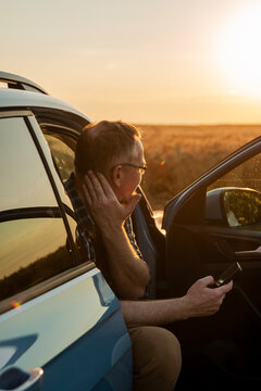 Middle-aged Caucasian farmer sitting in car with open door using smartphone at sunset. Smart farming concept.