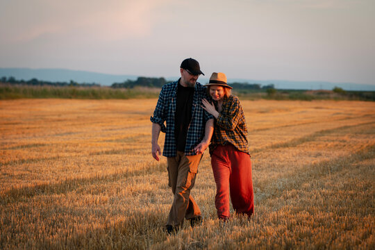 Happy middle aged farmer couple hugging and walking together through a harvested field at sunset