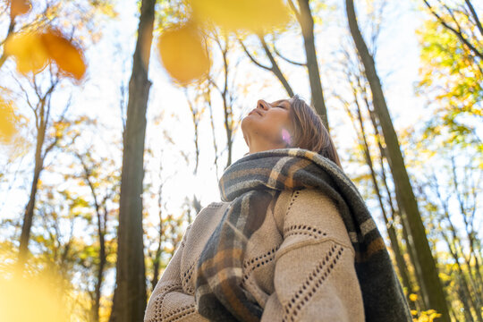 Mature Caucasian woman in warm sweater and scarf enjoying fresh air in autumn forest. Mental health and healthy aging concept