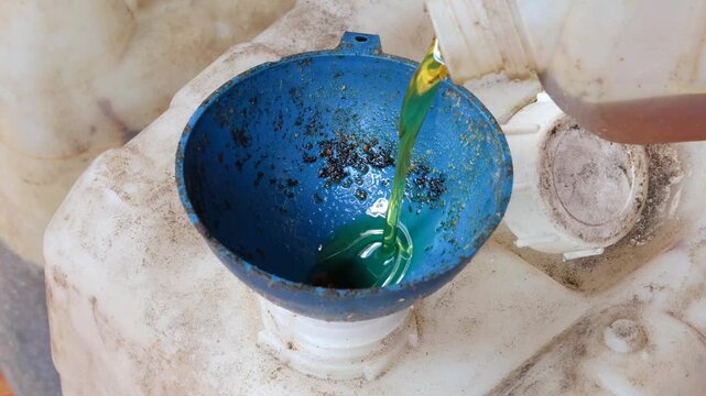 Close-up of an Asian man's hand pouring used cooking oil from a small jerry can into a larger plastic container using a funnel for household waste collection, recycling, and proper disposal.
