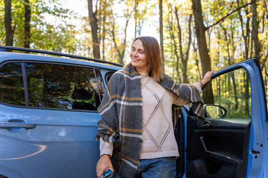 Cheerful mature woman wrapped in plaid blanket taking a break near vehicle, enjoying autumn sunshine. Healthy aging concept