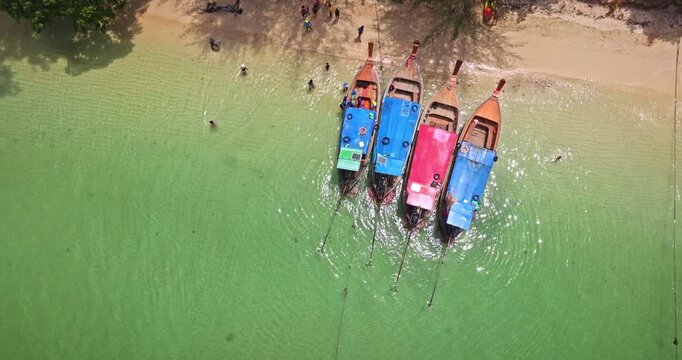 Four colorful long tail boats mooring on a sandy tropical beach, tourists swimming and relaxing in the clear turquoise waters of Koh Lanta, summer vacation and relax in Thailand. Aerial top down view