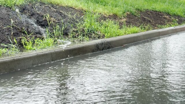 Storm water flows from a grassy bank into a flooded gutter with a steady splashing sound. Nearby cars drive through puddles creating a heavy white noise hum in the air.