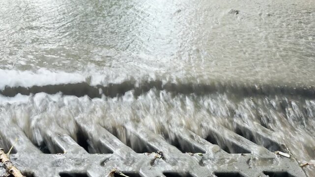 Turbid rainwater surges rapidly into a heavy metal storm drain. The fast movement creates a continuous roaring and splashing sound against the iron bars.