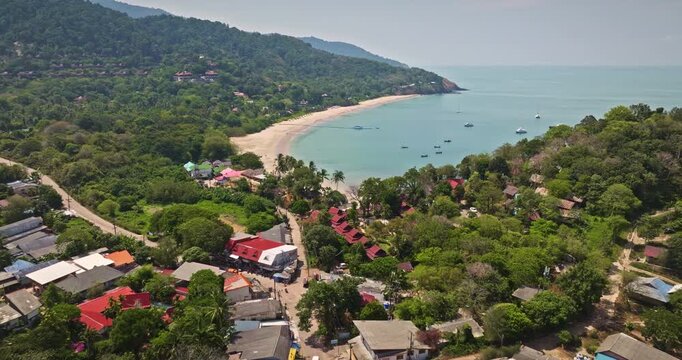 Aerial fight over Koh Lanta village buildings and lush green tropical trees line a winding road leading to a sandy beach and turquoise Andaman Sea bay with boats, surrounded by hills. Travel Thailand