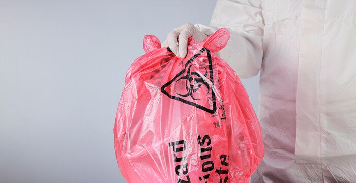 Cautious healthcare worker handles biohazard bag during clinical waste management wearing protective glove and suit against white background