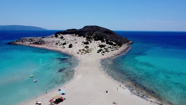 Drone footage of the iconic sandbar connecting the small islet at Simos Beach, Elafonisos, Greece, with turquoise water and white sand on a sunny summer day.