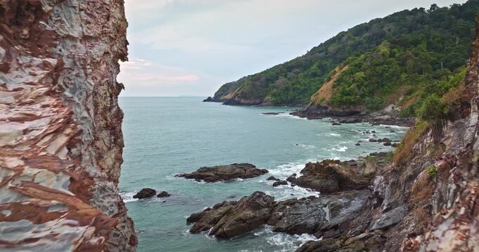 Drone fly in rock formations hole creating a natural frame. Showcasing the turquoise Andaman Sea meeting lush green coastline under a soft sky in Mu Ko Lanta National Park. Thailand. Aerial landscape
