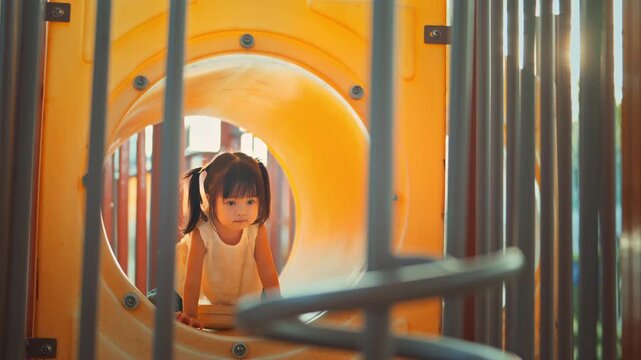 Cute Asian toddler girl crawling out of yellow tunnel playground sunset, active little kid playing outdoors in park for early childhood development, family leisure, outdoor activity and education