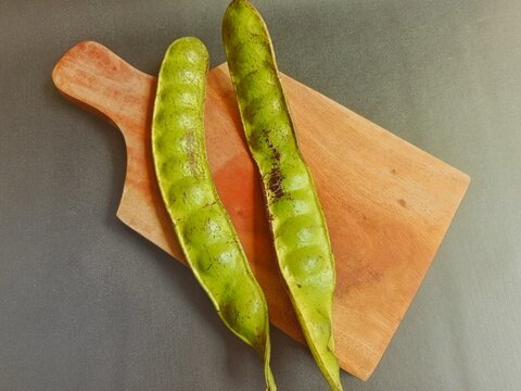 Fresh green petai or stink beans (Parkia speciosa) pods resting on a wooden cutting board against a dark rustic background.