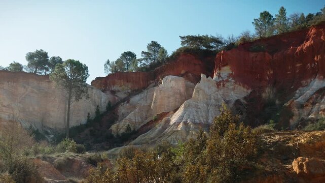 Detail of the large rock face in the expansive ochre rock landscape of Rustrel, France. The rugged red rocks are covered with trees and vegetation and are bathed in the morning sun.